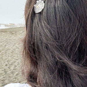 May include: A woman with long dark hair wearing a white shirt with a seashell and starfish hair clip.