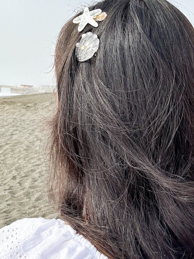 May include: A woman with long dark hair wearing a white shirt with a seashell and starfish hair clip.
