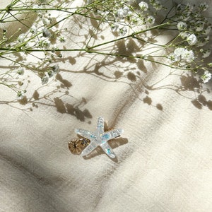 May include: A clear iridescent starfish hair clip with a gold accent. The clip is on a white fabric background with white flowers and their shadows.
