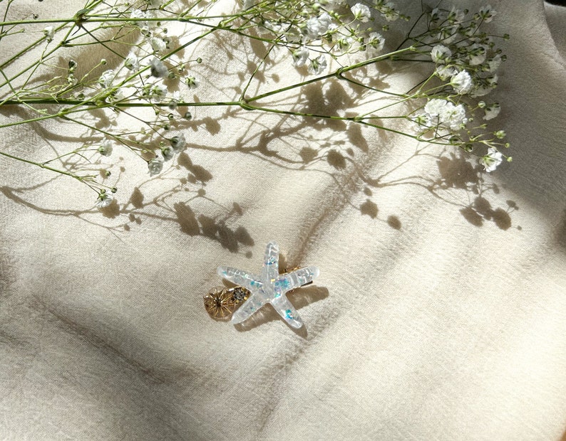 May include: A clear iridescent starfish hair clip with a gold accent. The clip is on a white fabric background with white flowers and their shadows.