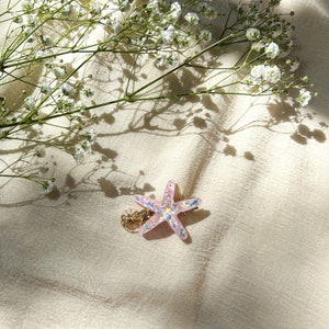 May include: A pink glitter starfish hair clip with a gold accent. The clip is on a white fabric background.
