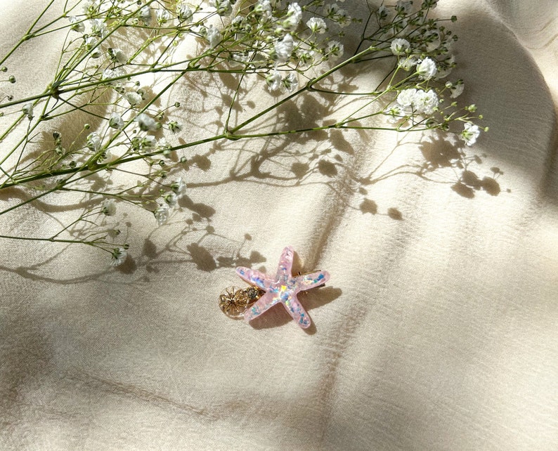 May include: A pink glitter starfish hair clip with a gold accent. The clip is on a white fabric background.