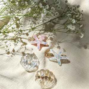 May include: Four hair clips with a beach theme. Two are clear plastic seashells with gold glitter and pearl accents. Two are starfishes, one is pink and one is clear with gold glitter.