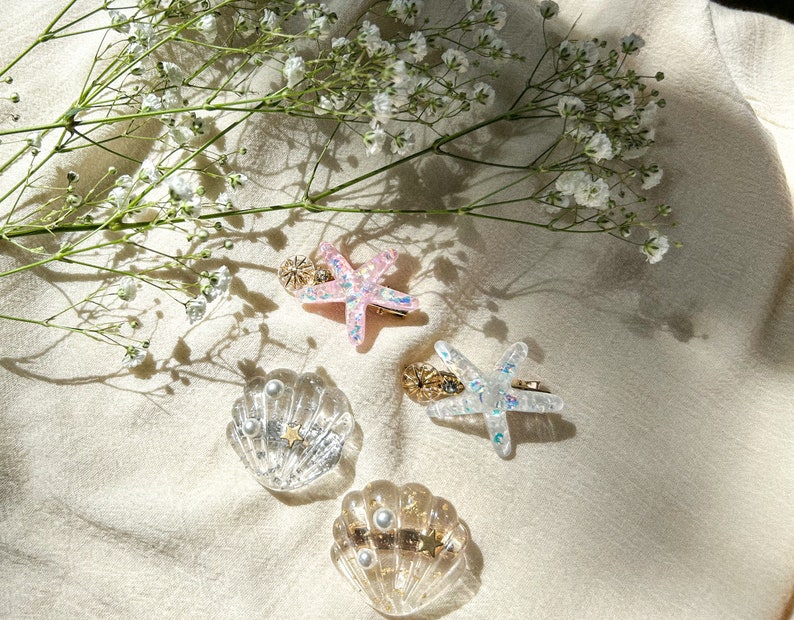 May include: Four hair clips with a beach theme. Two are clear plastic seashells with gold glitter and pearl accents. Two are starfishes, one is pink and one is clear with gold glitter.