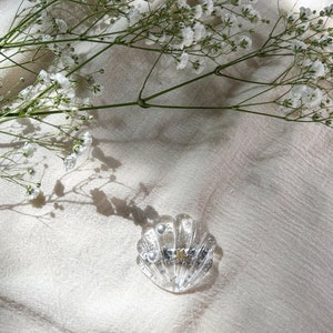 May include: A clear glass hair clip shaped like a seashell with a gold star and pearl accents. The clip is on a white fabric background.