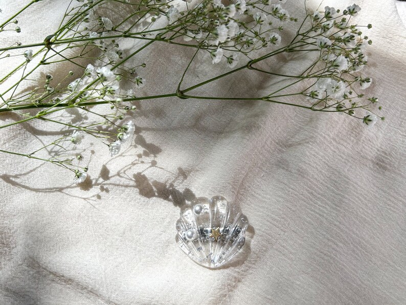 May include: A clear glass hair clip shaped like a seashell with a gold star and pearl accents. The clip is on a white fabric background.