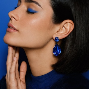 May include: A woman with dark hair and blue eyeliner wears large, teardrop-shaped blue earrings. The earrings are set in gold and dangle from the earlobes. The woman is wearing a dark blue sweater against a blue background.