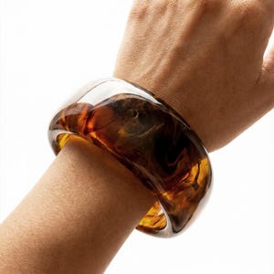 May include: A chunky, translucent brown and amber-colored bangle bracelet. The bracelet is worn on a wrist and has a marbled appearance. The jewelry is made of a glossy material and has a rounded shape.