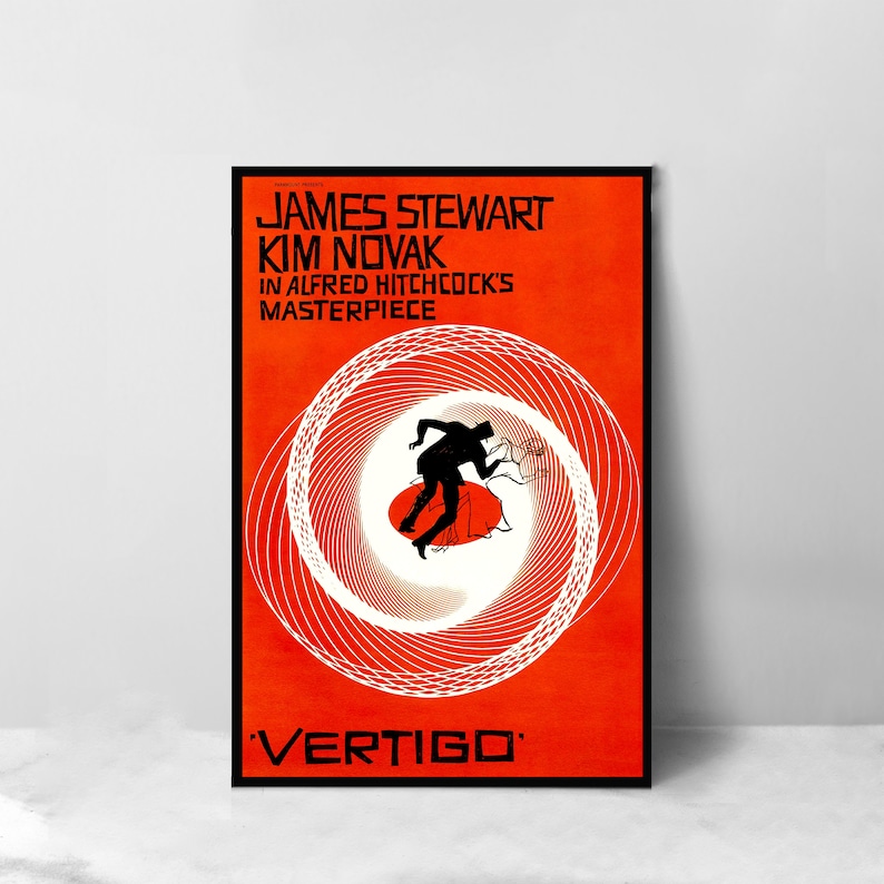 May include: A framed movie poster for Alfred Hitchcock's "Vertigo" featuring James Stewart and Kim Novak. The poster has a vibrant orange background with a white spiral design and a silhouette of a man falling. The title "Vertigo" is at the bottom.
