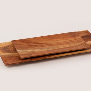 May include: Two wooden serving trays, one stacked on top of the other. The top tray is smaller and has a light brown color. The bottom tray is larger and has a darker brown color. Both trays have a natural wood grain pattern.