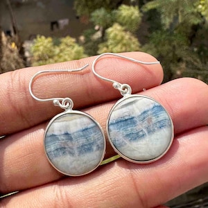 May include: A pair of silver earrings with round, marbled blue and white stone pendants. The earrings have a simple hook design. The stone's pattern resembles a landscape with layered blue and white hues.