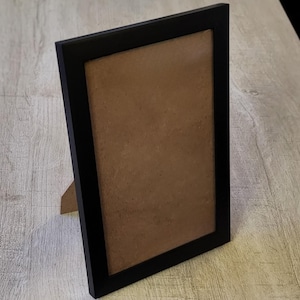 Può includere: Una cornice rettangolare nera con un retro marrone, in piedi. La cornice ha un design semplice e moderno ed è supportata da un piccolo cavalletto sul retro. La cornice è esposta su una superficie di legno di colore chiaro.