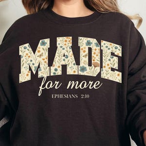 Può includere: Una felpa marrone scuro con le parole "MADE for more" in un design floreale. Il testo "Ephesians 2:10" è sotto il testo principale. La felpa è indossata da una persona.