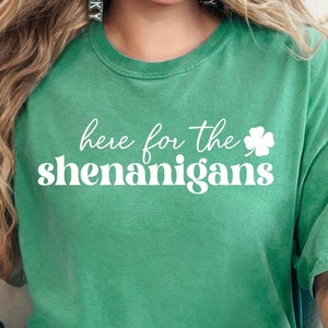 May include: A woman wearing a green t-shirt with the text "here for the shenanigans" and a white shamrock. She is sitting on a couch with a green shamrock pillow.