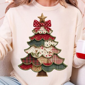 Può includere: Felpa color crema con un design a forma di albero di Natale. L'albero è composto da toppe di tessuto a strati in rosso, verde e beige con una stella dorata e un fiocco rosso in cima. È visibile anche una tazza rossa e bianca con un omino di pan di zenzero.