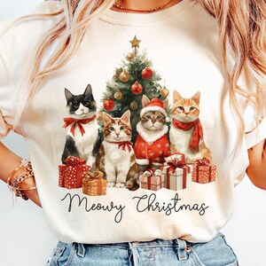 Può includere: T-shirt color crema con un design natalizio. La grafica presenta quattro gatti, un albero di Natale decorato e dei regali. I gatti indossano accessori festivi e il testo "Meowy Christmas" è visualizzato sotto l'immagine.