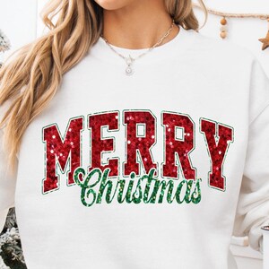 Può includere: Felpa bianca con una grafica "Buon Natale" in glitter rosso. La felpa è indossata da una persona seduta su una coperta bianca con un albero di Natale sullo sfondo.