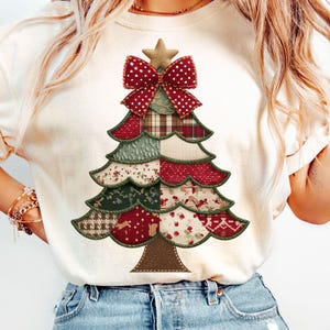 Può includere: T-shirt color crema con un design a forma di albero di Natale. L'albero è composto da vari modelli di tessuto in rosso, verde e bianco, sormontato da un fiocco rosso e una stella. Il design è centrato sulla maglietta.
