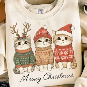 Può includere: Felpa color crema con tre gatti dei cartoni animati con maglioni e cappelli natalizi. I gatti indossano maglioni verdi, marroni e rossi con disegni festivi. Il testo "Meowy Christmas" è stampato sotto.