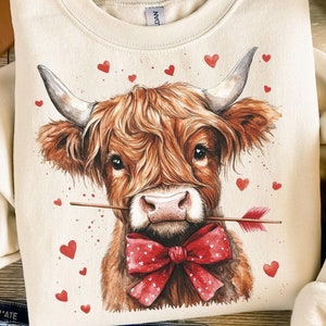 Pu&ograve; includere: Una felpa color crema con una grafica di una mucca Highland marrone e bianca con un papillon rosso e una freccia nel naso. La mucca &egrave; circondata da cuori rossi.