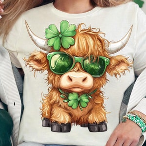 May include: A white long-sleeved shirt with a cartoon illustration of a brown Highland cow wearing green sunglasses and a green shamrock headband. The cow is looking directly at the viewer.