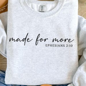 Può includere: Felpa grigio chiaro con la scritta "made for more" e "Ephesians 2:10" in corsivo nero. La felpa è ripiegata sopra jeans blu, con un cappello marrone e sandali sullo sfondo. L'immagine include anche icone di tipo di file SVG, PNG, EPS e DXF.