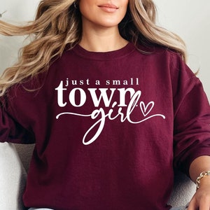 May include: A maroon sweatshirt with the text "just a small town girl" in white lettering with a small heart. The model is wearing the sweatshirt with blue jeans.