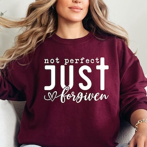 May include: A maroon sweatshirt with the text "not perfect JUST forgiven" in white lettering. A white heart and a white cross are also included in the design.