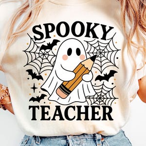 May include: Cream-colored t-shirt featuring a Halloween-themed graphic. The design includes a cartoon ghost holding a pencil, surrounded by a spiderweb, bats, and the words "Spooky Teacher". The shirt is paired with blue jeans.