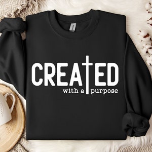 Può includere: Felpa girocollo nera con la scritta bianca "CREATED with a purpose". La lettera "T" di "CREATED" è sostituita da una croce. La felpa è presentata su un vassoio di legno, insieme a una tazza bianca e steli di cotone.