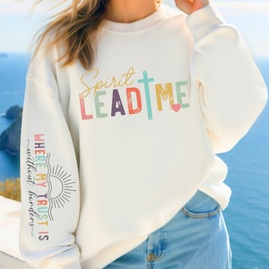 Puede incluir: Sudadera blanca con el texto "Spirit Lead Me" en colores arcoíris con una cruz y un corazón. La manga izquierda tiene el texto "Where my trust is without borders" en colores arcoíris con un gráfico de sol.
