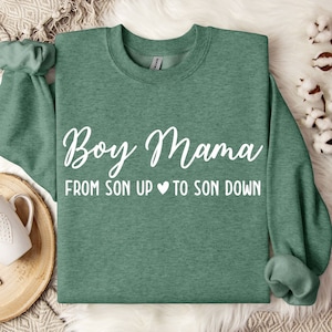 Pode incluir: Uma camisa de moletom verde com o texto "Boy Mama From Son Up to Son Down" em letras brancas. A camisa de moletom está sobre uma superfície branca e felpuda.