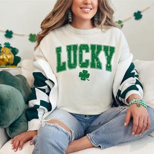 May include: A white sweatshirt with green text that says "LUCKY" and a green four-leaf clover. The sweatshirt has a white and green striped pattern on the sleeves.