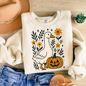 May include: Cream-colored sweatshirt featuring a whimsical goose design with a jack-o'-lantern and floral accents. The goose has an orange beak and feet. The design includes black leaves, orange flowers, and stars. The sweatshirt is paired with blue jeans and a tan hat.