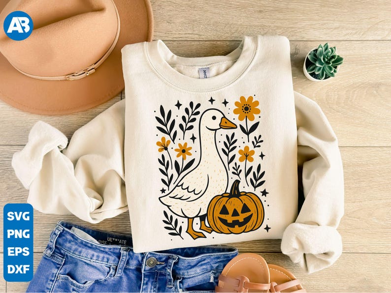 May include: Cream-colored sweatshirt featuring a whimsical goose design with a jack-o'-lantern and floral accents. The goose has an orange beak and feet. The design includes black leaves, orange flowers, and stars. The sweatshirt is paired with blue jeans and a tan hat.