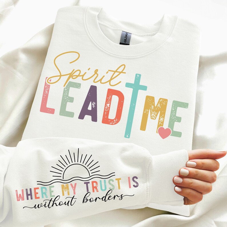 K&ouml;nnte beinhalten: Wei&szlig;er Pullover mit dem Text "Spirit Lead Me" in bunten Buchstaben mit einem Kreuz und einem Herzen. Der untere Teil des Pullovers hat den Text "Where my trust is without borders" mit einer Sonne und Wellengrafik.