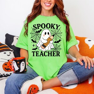 May include: A lime green t-shirt with the words "Spooky Teacher" and a graphic of a ghost holding a pencil. The shirt is paired with blue jeans. A black mug with white ghost designs is visible.