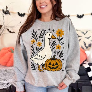 May include: Gray sweatshirt featuring a white goose with a pumpkin and floral design. The goose has an orange beak and feet. The pumpkin has a jack-o'-lantern face. The design includes yellow flowers and black leaves, perfect for Halloween.