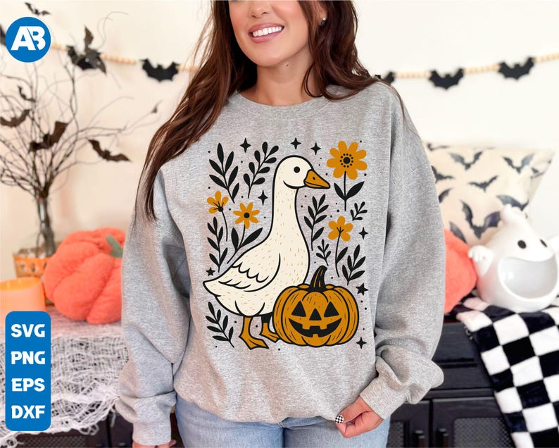 May include: Gray sweatshirt featuring a white goose with a pumpkin and floral design. The goose has an orange beak and feet. The pumpkin has a jack-o'-lantern face. The design includes yellow flowers and black leaves, perfect for Halloween.