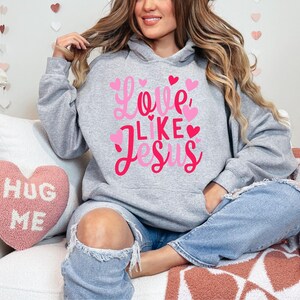 May include: A gray hoodie with the text "Love Like Jesus" in pink lettering and pink hearts. The model is wearing the hoodie with blue jeans.