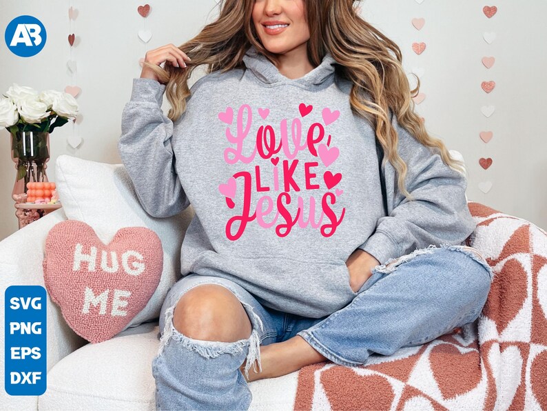 May include: A gray hoodie with the text "Love Like Jesus" in pink lettering and pink hearts. The model is wearing the hoodie with blue jeans.