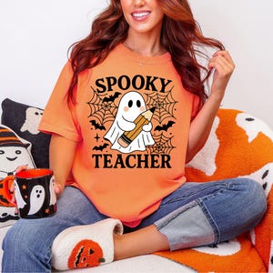 May include: Orange t-shirt with the words "Spooky Teacher" and a cartoon ghost holding a pencil. The design includes bats and a spiderweb. The person is wearing blue jeans and pumpkin-themed slippers.