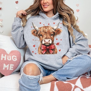 Pu&ograve; includere: Una felpa grigia con una mucca scozzese marrone e bianca che indossa un fiocco rosso e tiene una freccia, con cuori sullo sfondo. Un cuscino a forma di cuore rosa con il testo "Hug Me" &egrave; visibile.