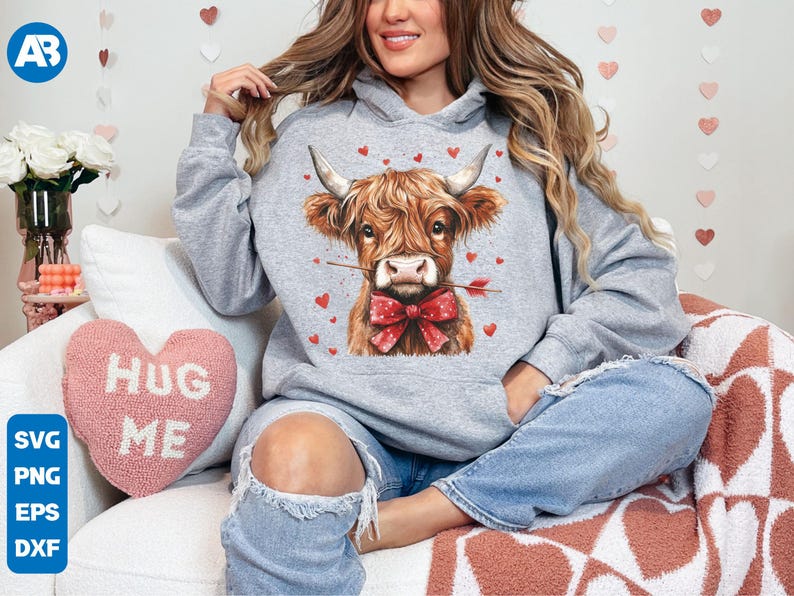 Pu&ograve; includere: Una felpa grigia con una mucca scozzese marrone e bianca che indossa un fiocco rosso e tiene una freccia, con cuori sullo sfondo. Un cuscino a forma di cuore rosa con il testo "Hug Me" &egrave; visibile.