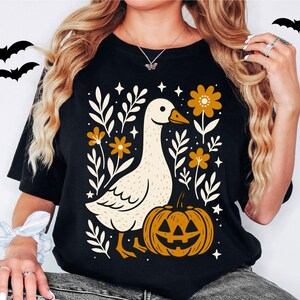 May include: Black t-shirt featuring a whimsical design with a goose, pumpkin, and floral elements. The goose is cream-colored with an orange beak, and the pumpkin has a carved face. The design includes orange flowers and white leaves, perfect for Halloween.