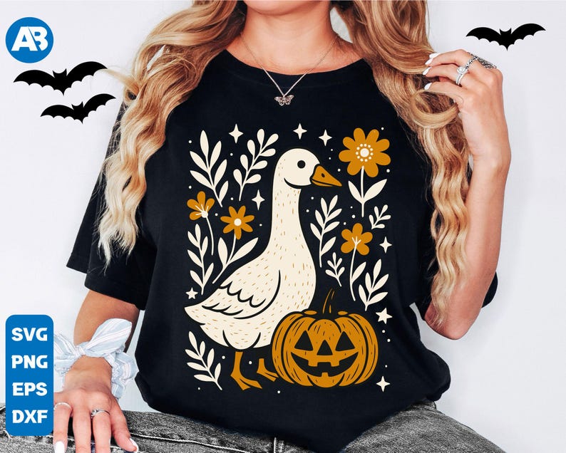 May include: Black t-shirt featuring a whimsical design with a goose, pumpkin, and floral elements. The goose is cream-colored with an orange beak, and the pumpkin has a carved face. The design includes orange flowers and white leaves, perfect for Halloween.
