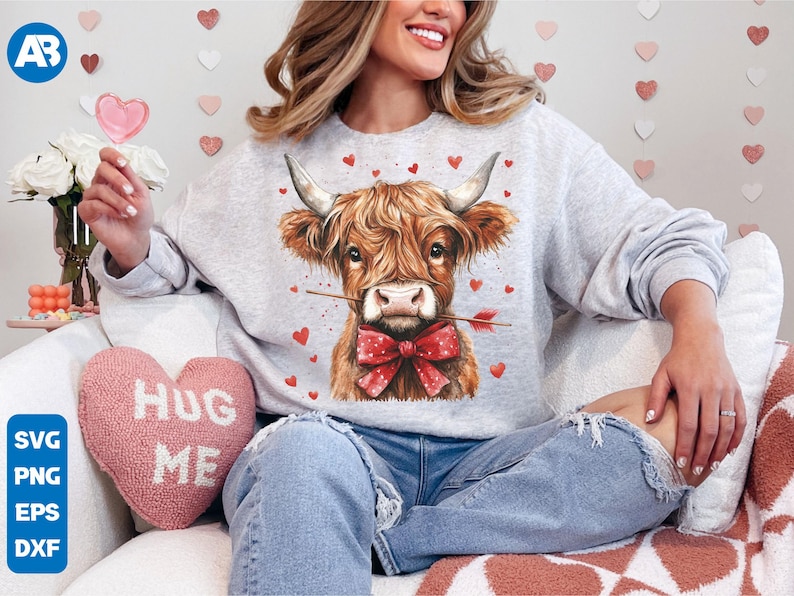 Pu&ograve; includere: Una felpa grigia con una mucca Highland marrone e bianca che indossa un papillon rosso e tiene una freccia in bocca. La mucca &egrave; circondata da cuori rossi. Il testo "Hug Me" &egrave; su un cuscino a forma di cuore rosa.