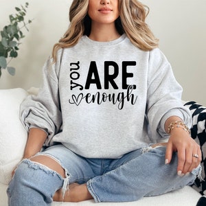 May include: A woman wearing a gray sweatshirt with the text "You Are Enough" in black lettering. The sweatshirt has a heart symbol before the word "enough". She is sitting on a white couch with a black and white checkered throw blanket.