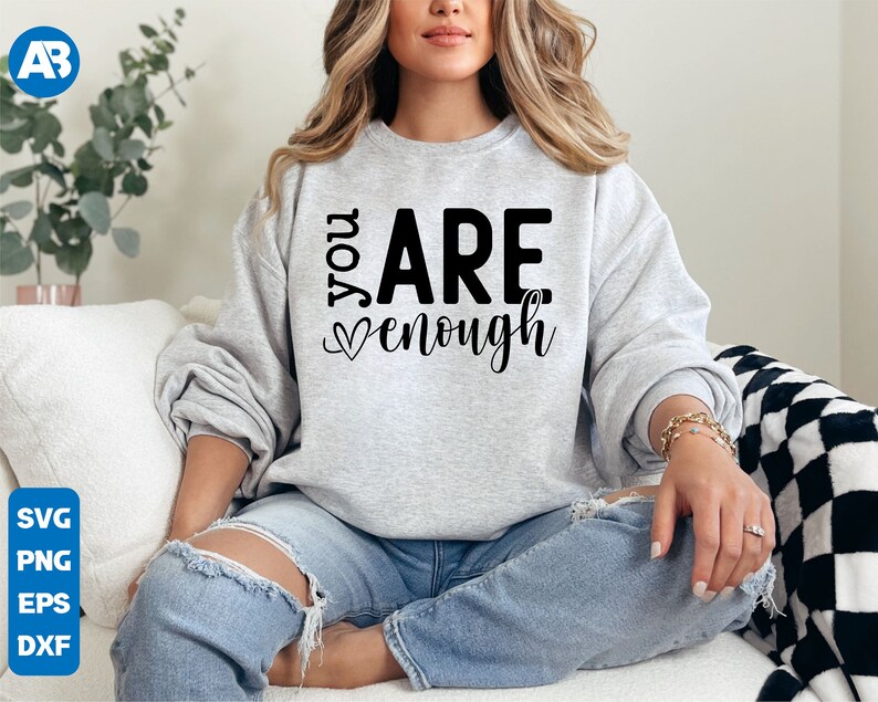 May include: A woman wearing a gray sweatshirt with the text "You Are Enough" in black lettering. The sweatshirt has a heart symbol before the word "enough". She is sitting on a white couch with a black and white checkered throw blanket.