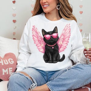 May include: A white sweatshirt with a black cat wearing pink angel wings and sunglasses. The cat is sitting on a white background. The text "Hug Me" is visible on a pink heart-shaped pillow in the background.